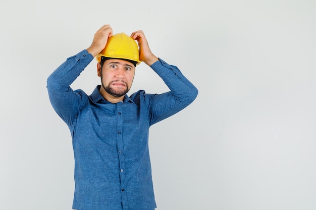 Junger arbeiter, der hände auf kopf in hemd, helm hält und hilflos schaut.