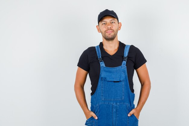 Junger Arbeiter, der Hände in den Taschen in der Uniform hält und fröhliche Vorderansicht schaut.