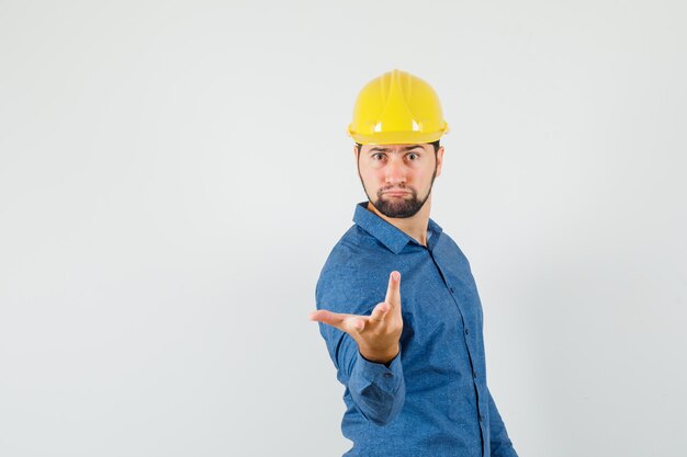 Junger Arbeiter, der Hand in verwirrter Geste in Hemd, Helm streckt