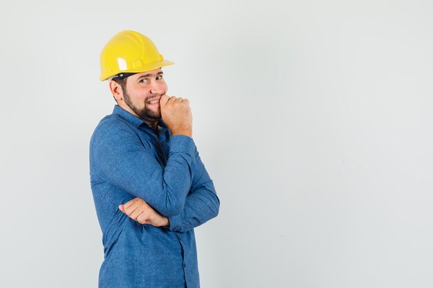 Junger Arbeiter, der in der denkenden Haltung im Hemd, im Helm steht und beschämt schaut