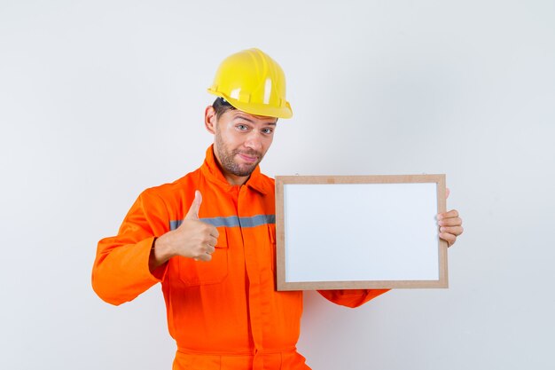 Junger Arbeiter, der leeren Rahmen hält, Daumen in Uniform, Helm zeigt und froh aussieht.