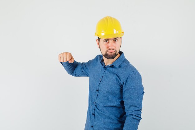 Junger Arbeiter, der mit Faust in Hemd, Helm droht und nervös schaut