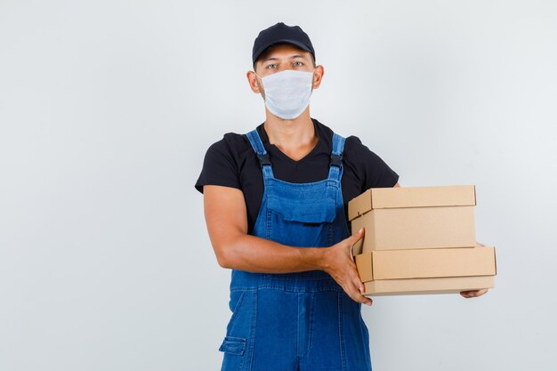 Junger Arbeiter, der Pappkartons in Uniform, Maskenvorderansicht hält.