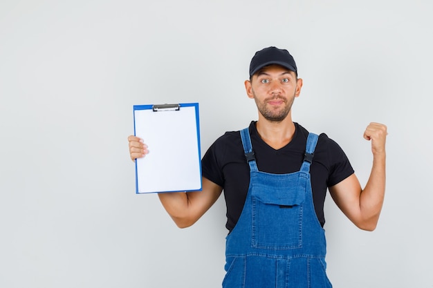 Kostenloses Foto junger arbeiter, der zurück zeigt und zwischenablage in der einheitlichen vorderansicht hält.