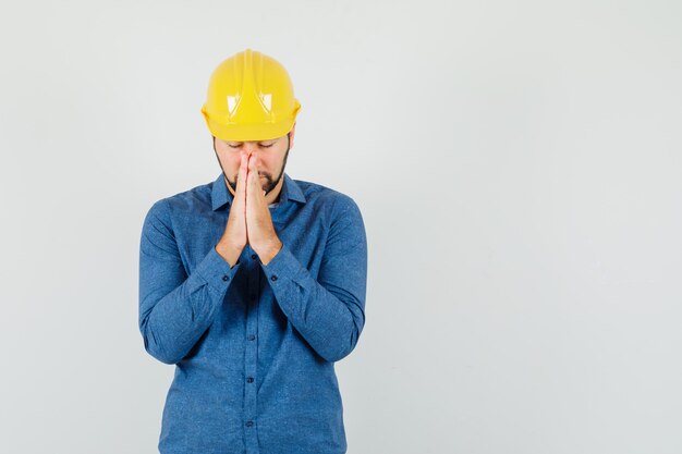 Junger Arbeiter im Hemd, Helm, der Hände in der Gebetsgeste hält und ruhig schaut