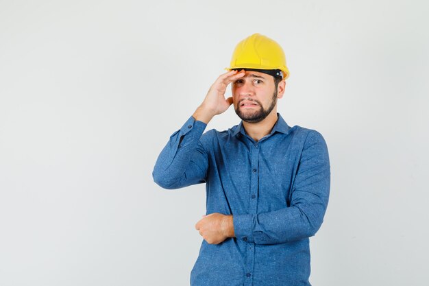 Junger Arbeiter im Hemd, Helm, der Hand über Gesicht hält und verlegen aussieht