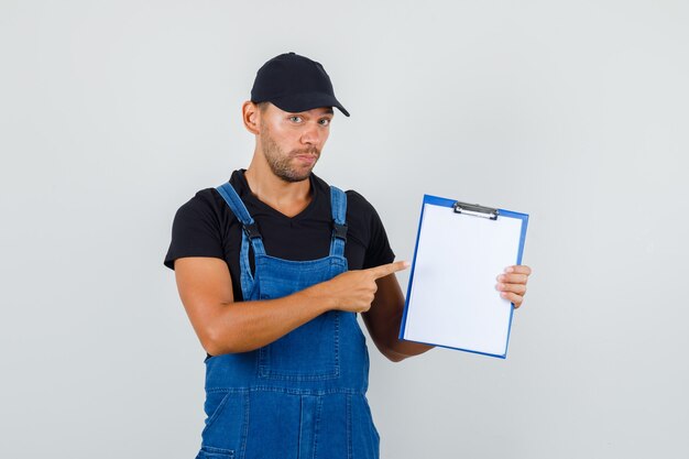 Junger Arbeiter in der Uniform, die auf Zwischenablage zeigt, Vorderansicht.