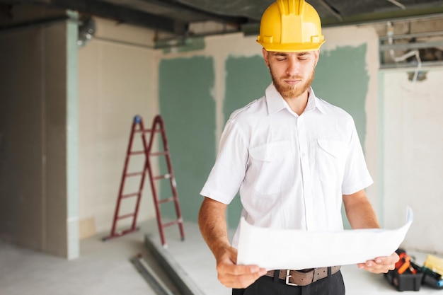 Junger Architekt in weißem Hemd und gelbem Helm blickt nachdenklich auf den Plan der neuen Wohnung mit Leiter im Hintergrund