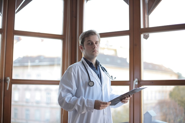 Junger Arzt, der in seinem Büro mit Fenstern auf dem Hintergrund steht