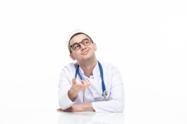 Junger Arzt der Vorderansicht im medizinischen Anzug, der hinter Schreibtisch sitzt