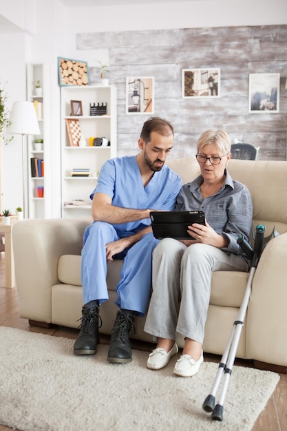 Junger Arzt im Pflegeheim hilft Seniorin mit Krücken, Tablet-Computer zu benutzen.