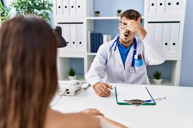 Junger Arzt mit Klient in der medizinischen Klinik, der schockiert späht, Gesicht und Augen mit der Hand bedeckt und mit verlegenem Ausdruck durch die Finger schaut.