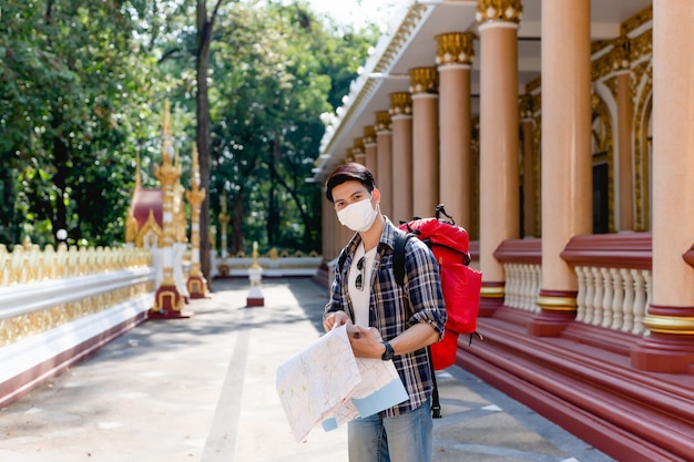 Junger asiatischer Backpacker-Mann in Gesichtsmaske, der die Richtung auf der Papierkarte in der Hand am schönen thailändischen Tempel steht und auf der Karte zeigt