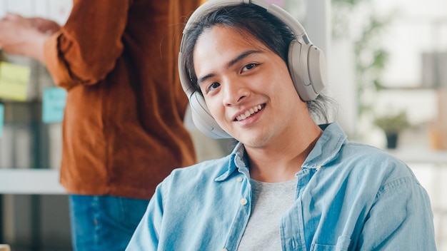 Junger asiatischer Designermann, der Musik auf Kopfhörern hört, Kamera betrachtet und im modernen Büro lächelt. Gruppe junger College-Studenten in schicker Freizeitkleidung auf dem Campus. Teamwork-Konzept für Mitarbeiter.