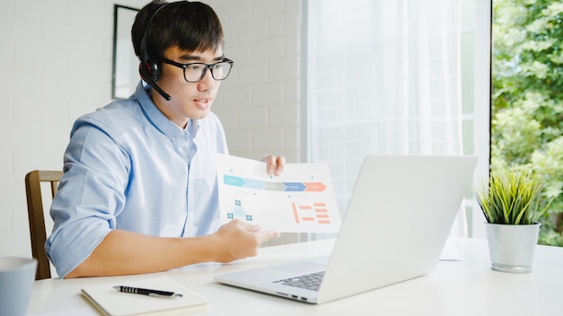 Junger asiatischer Geschäftsmann, der Laptop verwendet, spricht mit Kollegen über Plan in Videoanruf, während kluges Arbeiten von zu Hause im Wohnzimmer. Selbstisolation, soziale Distanzierung, Quarantäne zur Vorbeugung von Koronaviren.