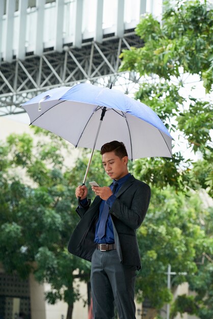 Junger asiatischer Geschäftsmann, der mit Regenschirm in der Straße steht und Smartphone verwendet