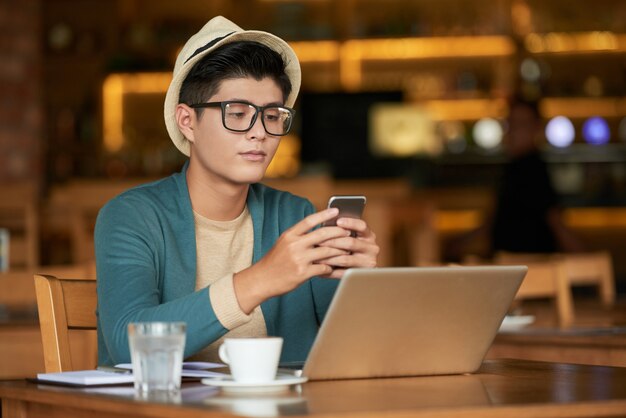 Junger asiatischer Hippie-Mann, der im Café mit Laptop sitzt und Smartphone verwendet