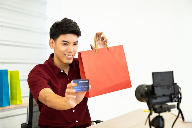 Junger asiatischer männlicher Online-Händler-Blogger, der mit Kamera Live-Video zeigt, zeigt Einkaufstasche und Kreditkarte Social Media Influencer Online-Shopping- und Zahlungskonzept
