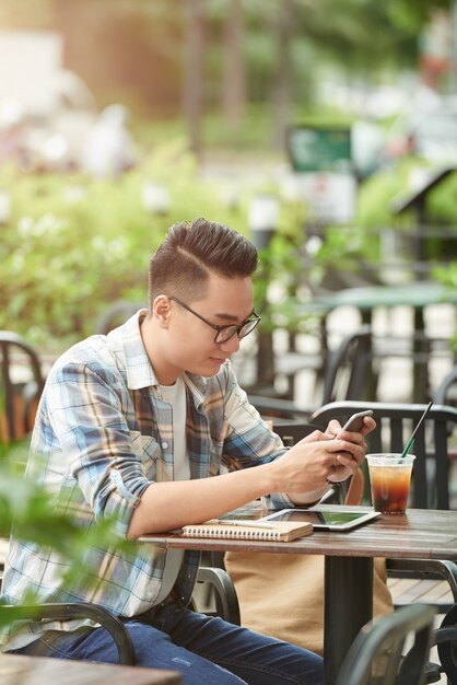 Junger asiatischer männlicher Student, der am Straßencafé sitzt und Smartphone verwendet