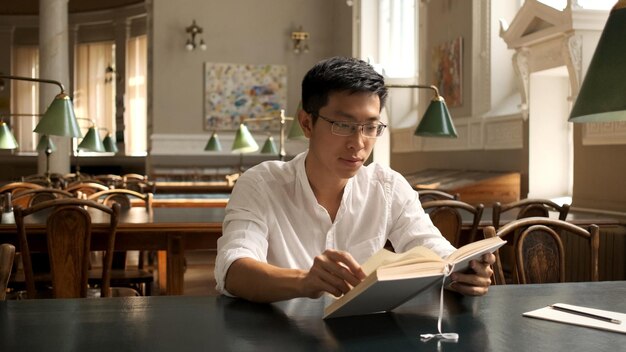 Junger asiatischer männlicher student, der intensiv mit lehrbuch studiert und sich auf prüfungen in der universitätsbibliothek vorbereitet