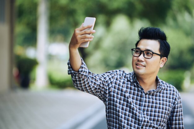 Junger asiatischer Mann in den Gläsern und im karierten Hemd, die selfie mit Smartphone nehmen