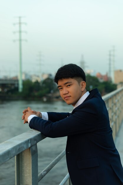 Junger asiatischer Mann in einem Anzug, der auf einer Brücke mit den Händen auf Geländern steht