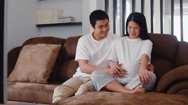 Junger asiatischer schwangerer Paarmann berühren seinen Fraubauch, der mit seinem Kind spricht. Die Mutter und Vati, die glücklich lächeln friedlich sich fühlen, während mach s gut Baby, die Schwangerschaft, die zu Hause auf Sofa im Wohnzimmer liegt.
