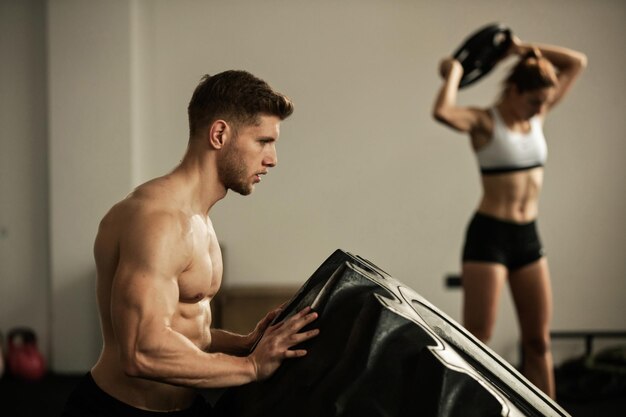 Junger athletischer Mann, der beim Cross-Training in einem Fitnessstudio Reifen-Flip-Übungen macht. Im Hintergrund arbeitet eine Frau mit Gewichten