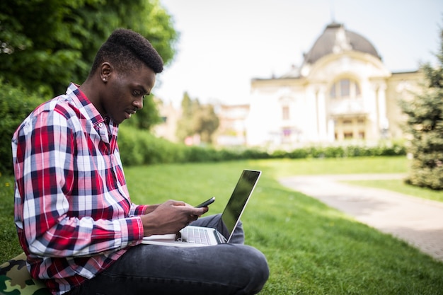 Junger attraktiver afroamerikanischer Geschäftsmann mit Laptop, der auf dem Gras sitzt und auf Handy im Park spricht