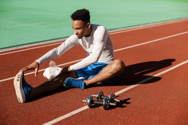 Kostenloses Foto junger attraktiver afroamerikanischer sportler, der sich auf der rennstrecke im stadtstadion ausdehnt