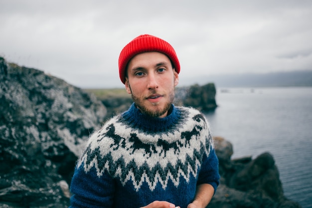 Junger attraktiver bärtiger tausendjähriger Mann im roten Fischer- oder Matrosenmützenhut und im traditionellen blauen Pullover der isländischen Verzierung