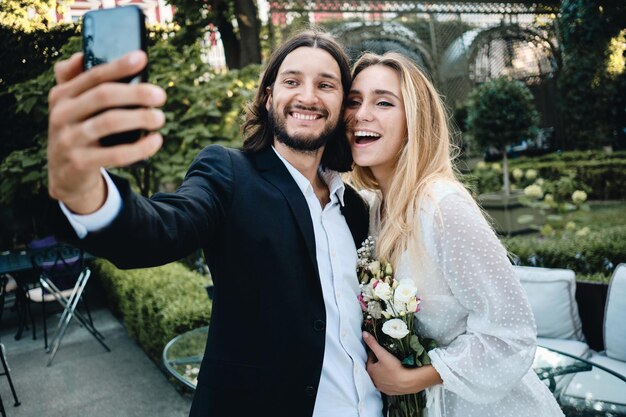 Junger attraktiver Bräutigam und Braut, die freudig zusammen Selfie auf dem Handy im schönen Garten des Restaurants machen