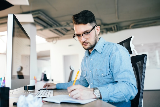 Junger attraktiver dunkelhaariger Mann in Glaslosigkeit arbeitet mit einem Computer und schreibt im Notizbuch im Büro. Er trägt ein blaues Hemd und einen Bart.