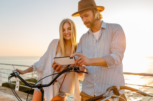 Junger attraktiver lächelnder glücklicher glücklicher Mann und Frau, die auf Fahrrädern mit Smartphone reisen, romantisches Paar am Meer bei Sonnenuntergang, Boho-Hipster-Stil-Outfit, Freunde, die Spaß zusammen haben