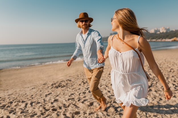 Junger attraktiver lächelnder glücklicher Mann im Hut und in der blonden Frau im weißen Kleid, das zusammen am Strand läuft