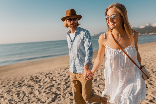 Junger attraktiver lächelnder glücklicher mann im hut und in der blonden frau im weißen kleid, das zusammen am strand läuft