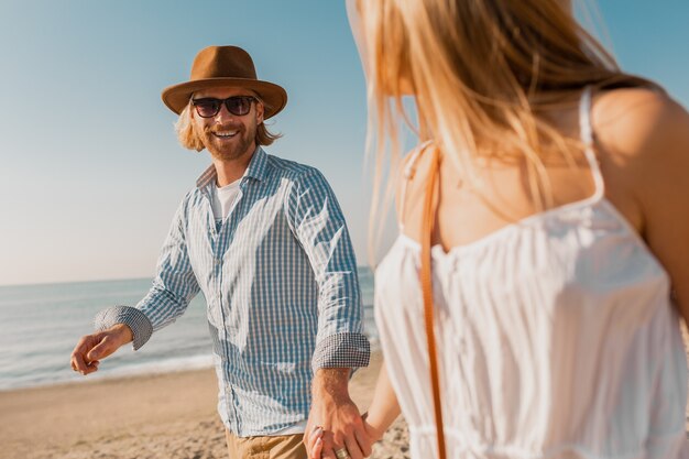 Junger attraktiver lächelnder glücklicher Mann im Hut und in der blonden Frau im weißen Kleid, das zusammen am Strand läuft