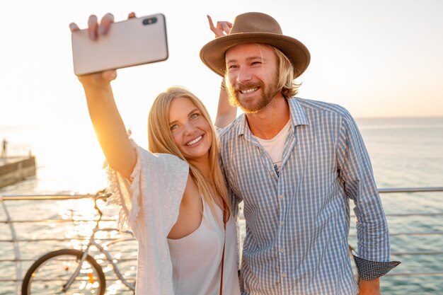 Junger attraktiver lächelnder glücklicher Mann und Frau, die auf Fahrrädern reisen, die Selfie-Foto auf Telefonkamera machen, romantisches Paar am Meer auf Sonnenuntergang, Boho-Hipster-Art-Outfit, Freunde, die Spaß zusammen haben