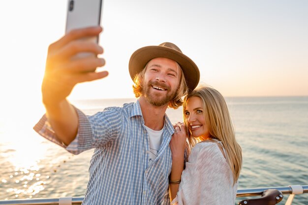 Junger attraktiver lächelnder glücklicher Mann und Frau, die auf Fahrrädern reisen, die Selfie-Foto auf Telefonkamera, romantisches Paar am Meer auf Sonnenuntergang, Boho-Hipster-Art-Outfit, Freunde haben, die Spaß zusammen haben