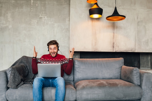 Junger attraktiver lächelnder Mann, der auf dem Sofa zu Hause im Winter mit überraschtem Gesichtsausdruck sitzt, roten Strickpullover trägt, am Laptop arbeitet, freiberuflich, emotional, schreiend, Kopfhörer hörend