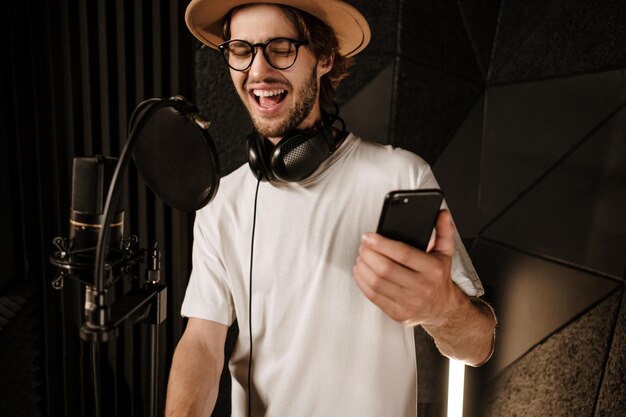 Junger attraktiver männlicher Sänger mit Smartphone singt emotional im modernen Tonstudio