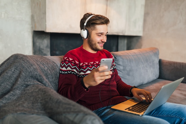 Junger attraktiver Mann auf Sofa zu Hause im Winter mit Smartphone in Kopfhörern, Musik hörend, roten Strickpullover tragend, am Laptop arbeitend, freiberuflich, lächelnd, glücklich, positiv, tippend