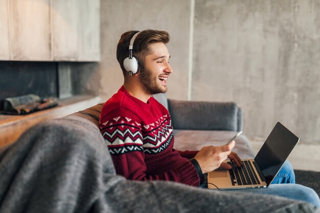 Junger attraktiver Mann auf Sofa zu Hause im Winter mit Smartphone in Kopfhörern, Musik hörend, roten Strickpullover tragend, am Laptop arbeitend, freiberuflich, lächelnd, glücklich, positiv