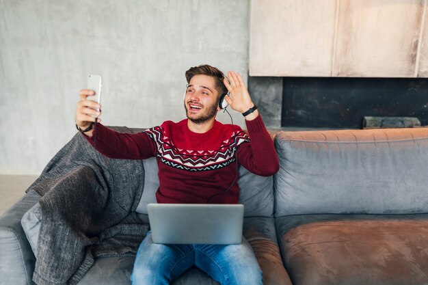 Junger attraktiver Mann auf Sofa zu Hause im Winter mit Smartphone in Kopfhörern, Musik hörend, tragender roter Strickpullover, Arbeiten am Laptop, Freiberufler