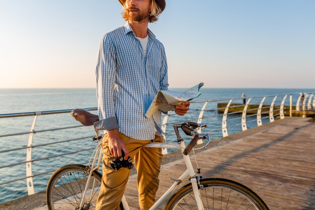 Junger attraktiver Mann, der auf Fahrrad durch See in den Sommerferien durch das Meer auf Sonnenuntergang, Boho-Hipster-Art-Outfit reist, hält Kartenbesichtigung, die Foto auf Kamera, gekleidet in Hemd und Hut hält
