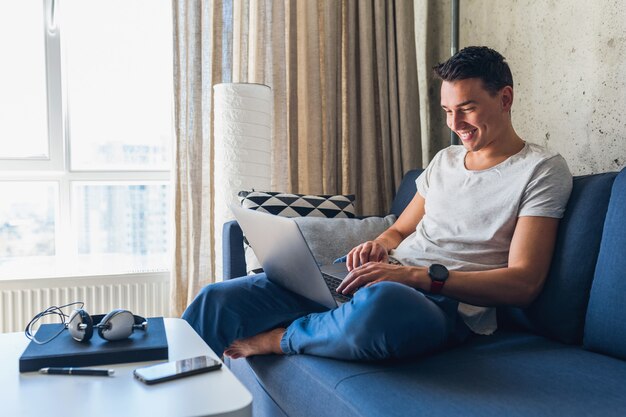Junger attraktiver Mann, der auf Sofa zu Hause sitzt und online am Laptop arbeitet, unter Verwendung des Internets