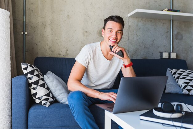 Junger attraktiver Mann, der auf Sofa zu Hause sitzt und online am Laptop arbeitet, unter Verwendung des Internets