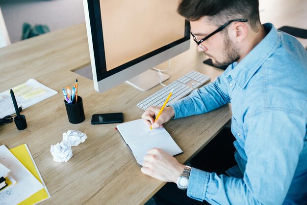 Junger attraktiver Mann mit Brille arbeitet an seinem Arbeitsplatz im Büro. Er trägt blaues Hemd. Er schreibt im Notizbuch. Sicht von oben.