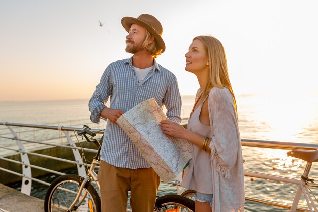 Junger attraktiver Mann und Frau, die auf Fahrrädern reisen, Karte und Besichtigung halten, romantisches Paar in den Sommerferien am Meer am Sonnenuntergang, Freunde, die Spaß zusammen haben