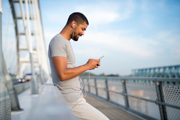 Junger attraktiver sportlicher Mann, der Telefon und Lächeln auf der Brücke verwendet
